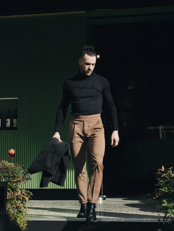 Stylish man in black turtleneck and beige pants walking outdoors in sunlight.