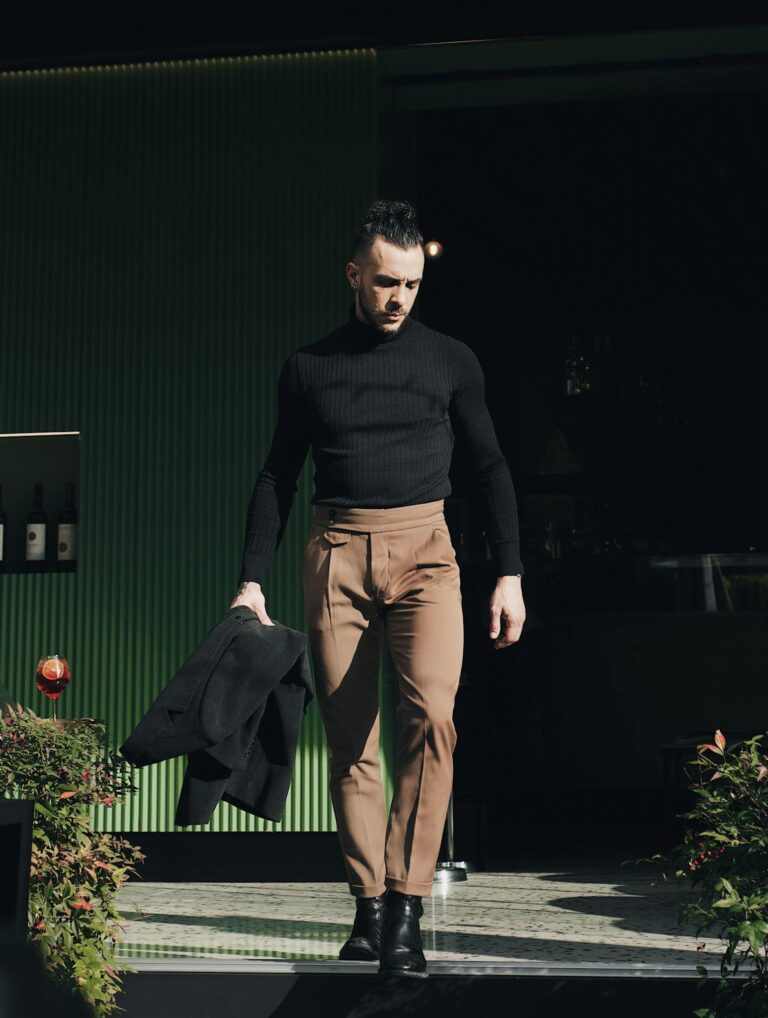 Stylish man in black turtleneck and beige pants walking outdoors in sunlight.